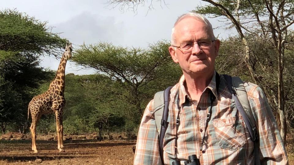 a giraffe in the background on the left looks at Douglas Cavener wearing a button-up and binoculars in the foreground on the right.